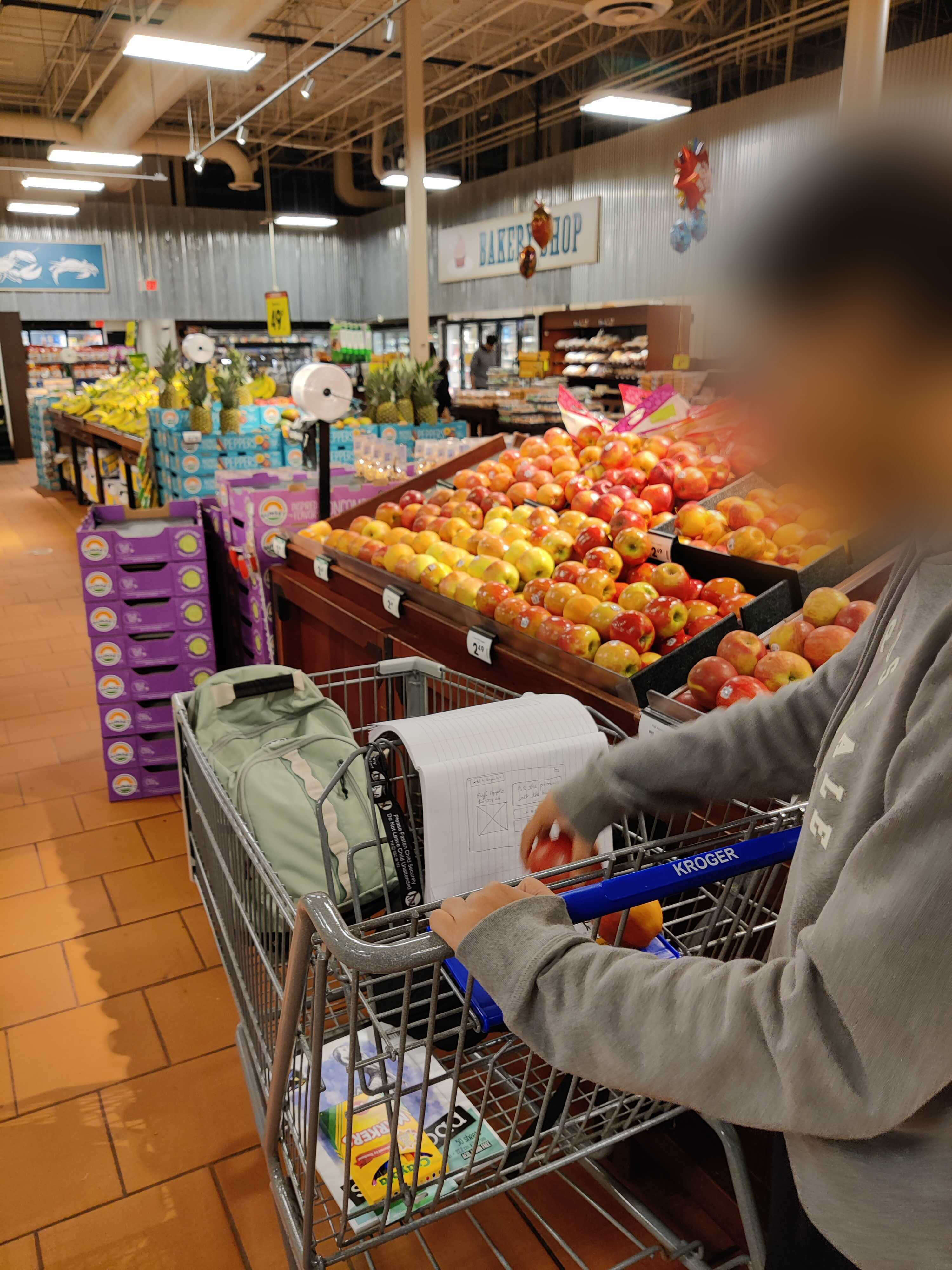 Shopper at a Kroger produce section adding fruit to a cart with a notebook propped up as the screen